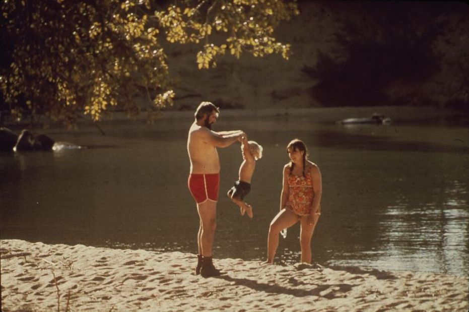 A faded 1970s colour snapshot: a father swings a toddler by the arms at the edge of a lake while the mother watches from the sand.